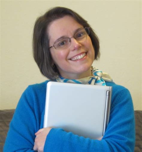 A woman smiling while holding a laptop close to her chest, wearing a blue sweater and a light-colored scarf.